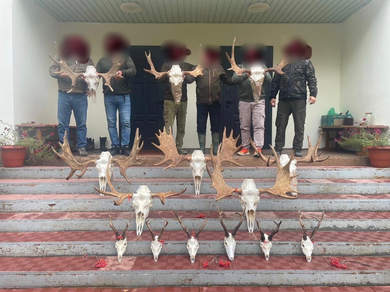 Six hunters behind a row of Siberian roe buck and moose trophies on lodge steps, Russia